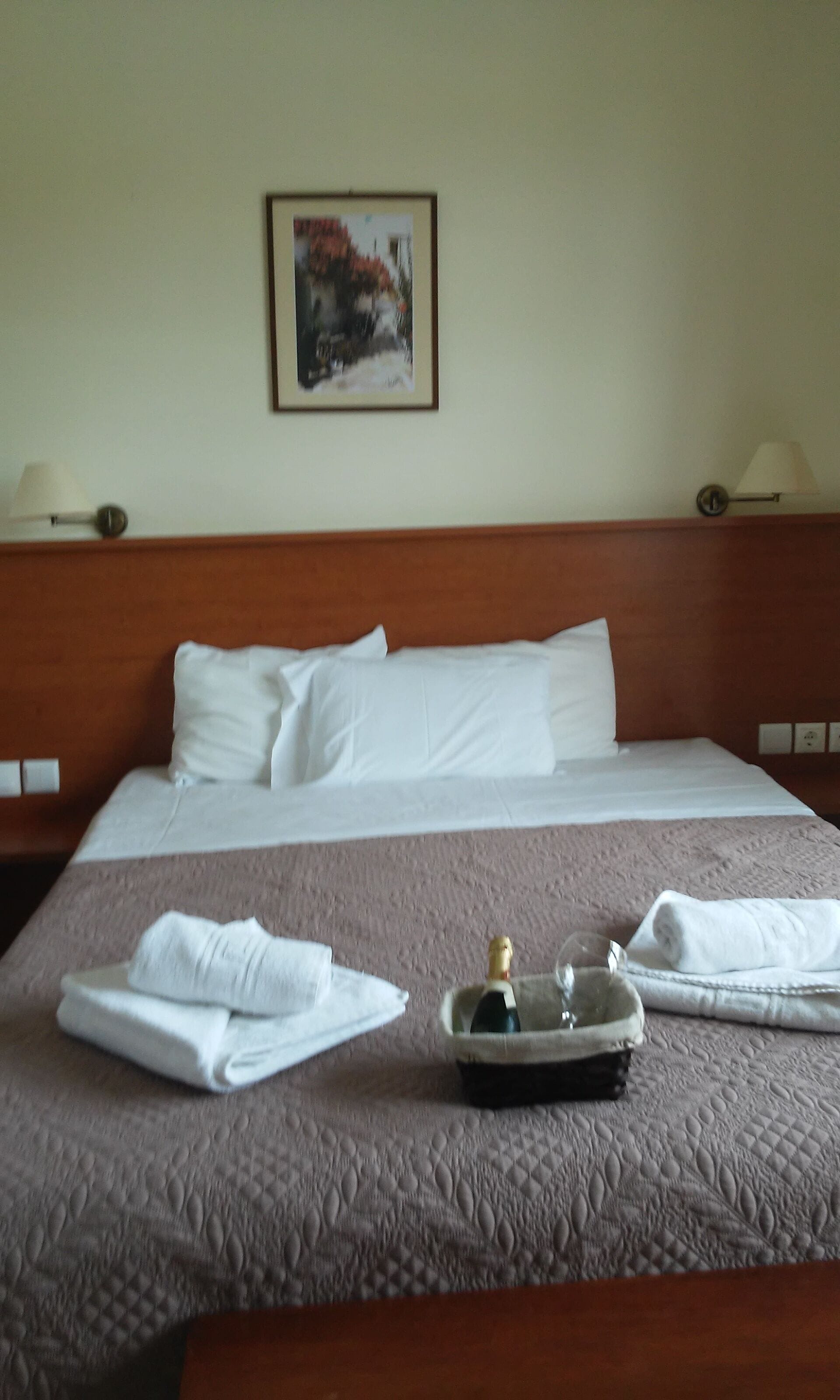 A neatly made bed with white linens, a bottle of champagne, and folded towels on a textured bedspread.