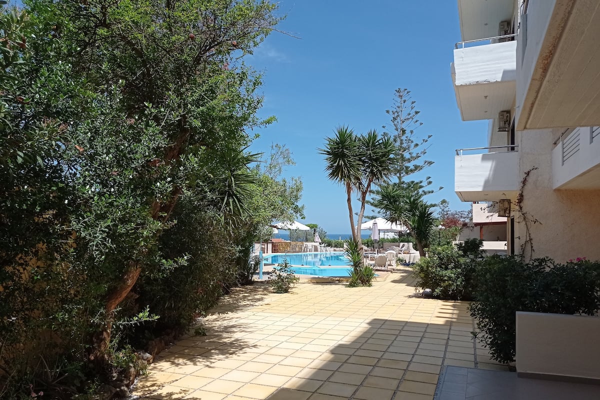 A pool area with tiled flooring, surrounded by greenery and a clear sky.