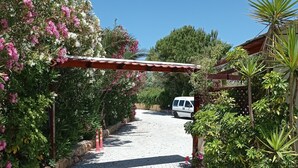 A paved pathway leading to a white van parked under a pergola with pink flowers. - Alexandros Studios (Chania)