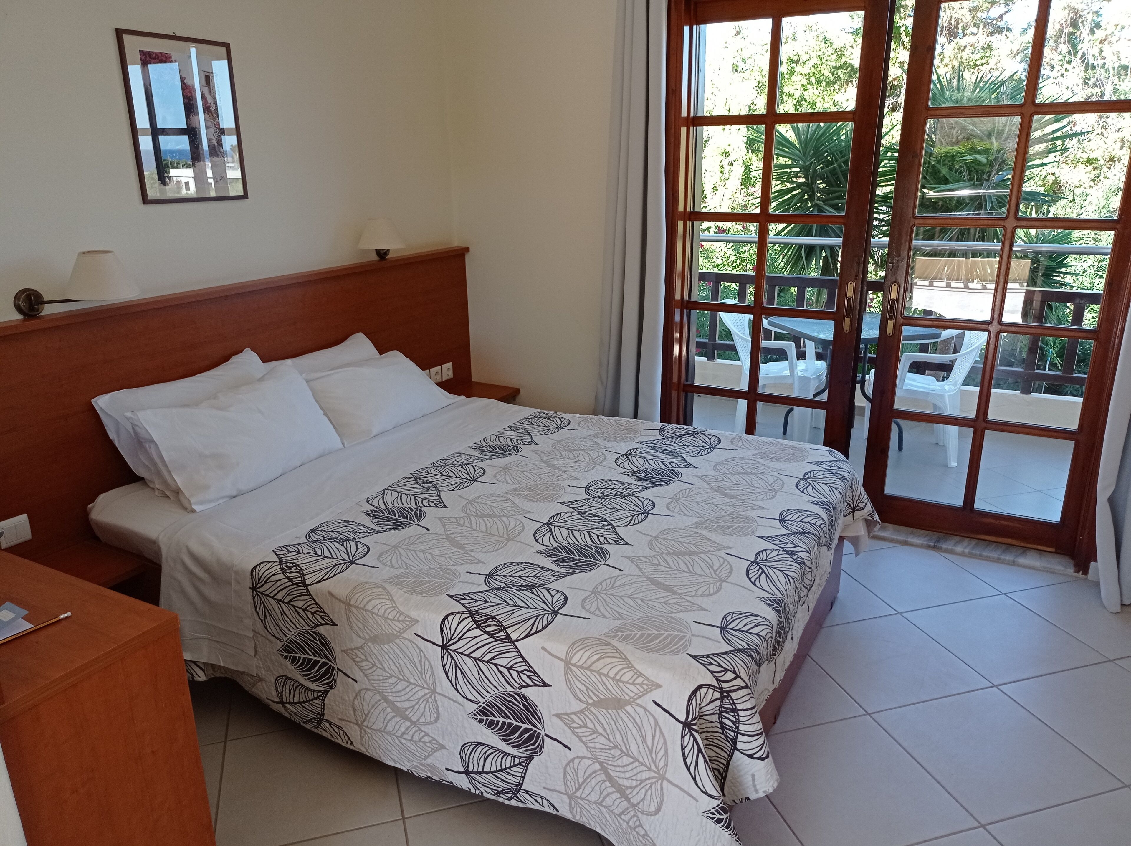 A neatly made bed with a leaf-patterned bedspread, a wooden headboard, and a view of an outdoor patio with tables and chairs through a large window.