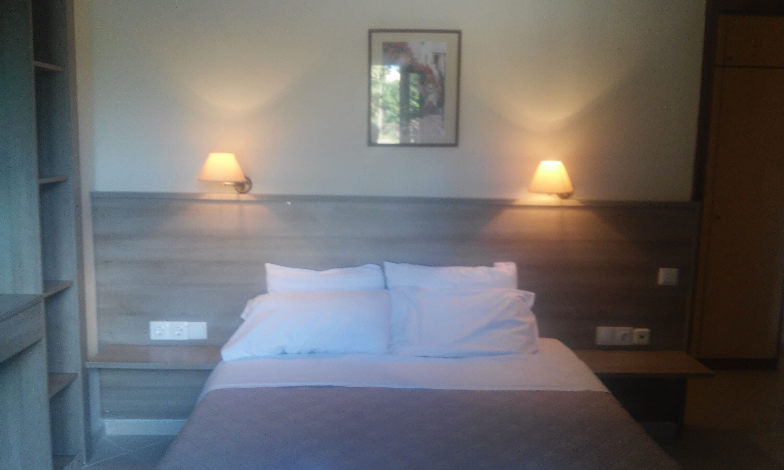 A hotel room with a bed, two wall-mounted lamps, and a framed picture on the wall.
