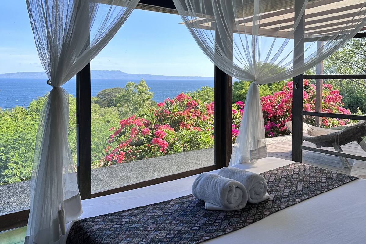Une chambre avec un grand lit, des rideaux blancs et une vue sur la mer et des fleurs roses.