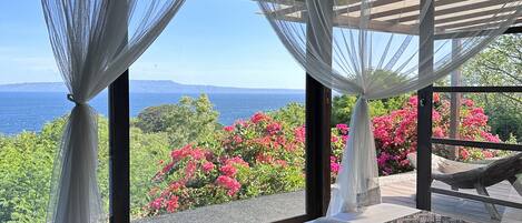 A bedroom with a large bed, white curtains, and a view of the sea and pink flowers.