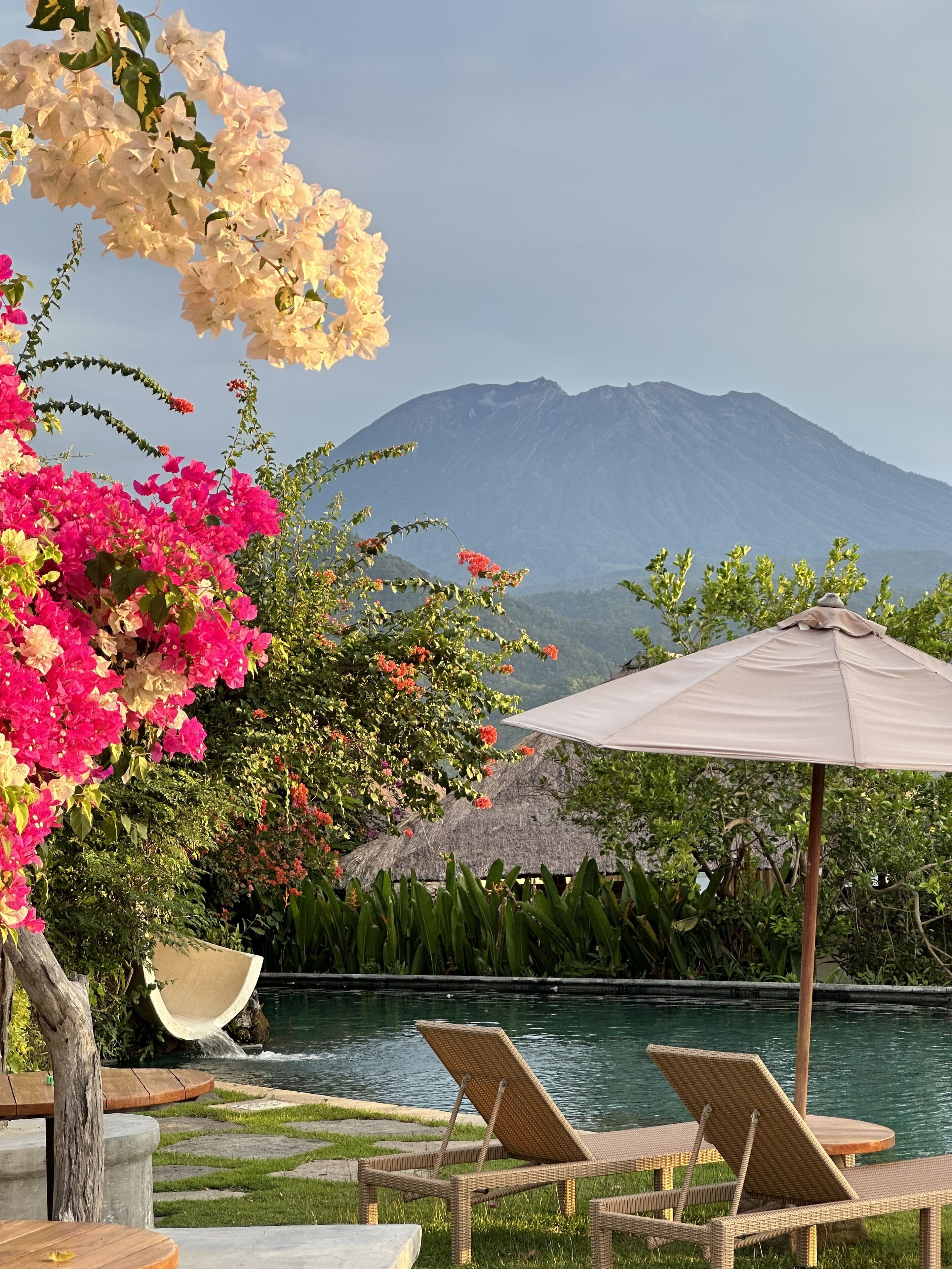 A poolside area with lounge chairs and an umbrella, surrounded by lush greenery and a mountain view.