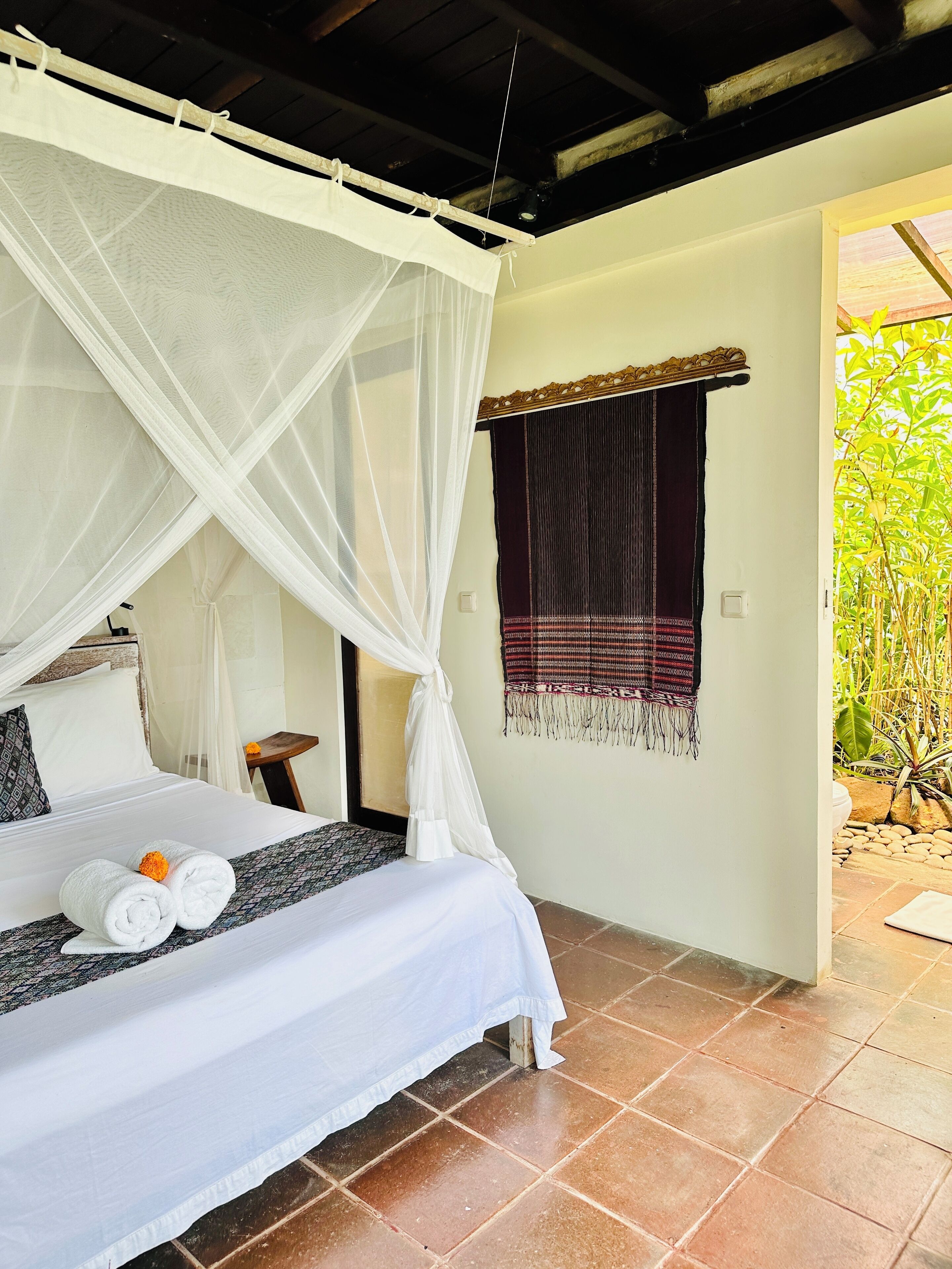 A bedroom with a bed, white curtains, a small table, and a view of greenery outside.