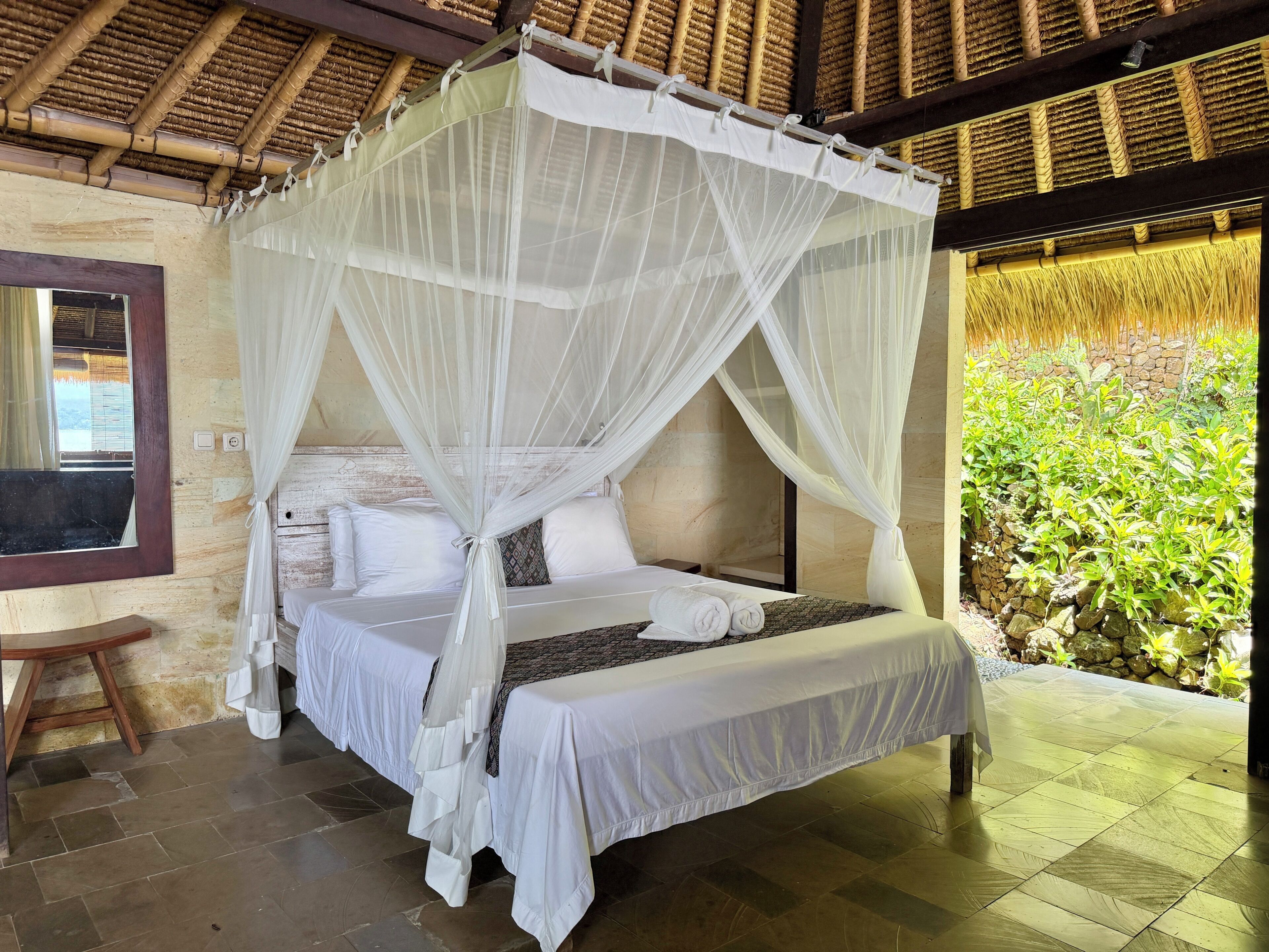 A bed with a canopy, a small table, and a view of greenery outside.