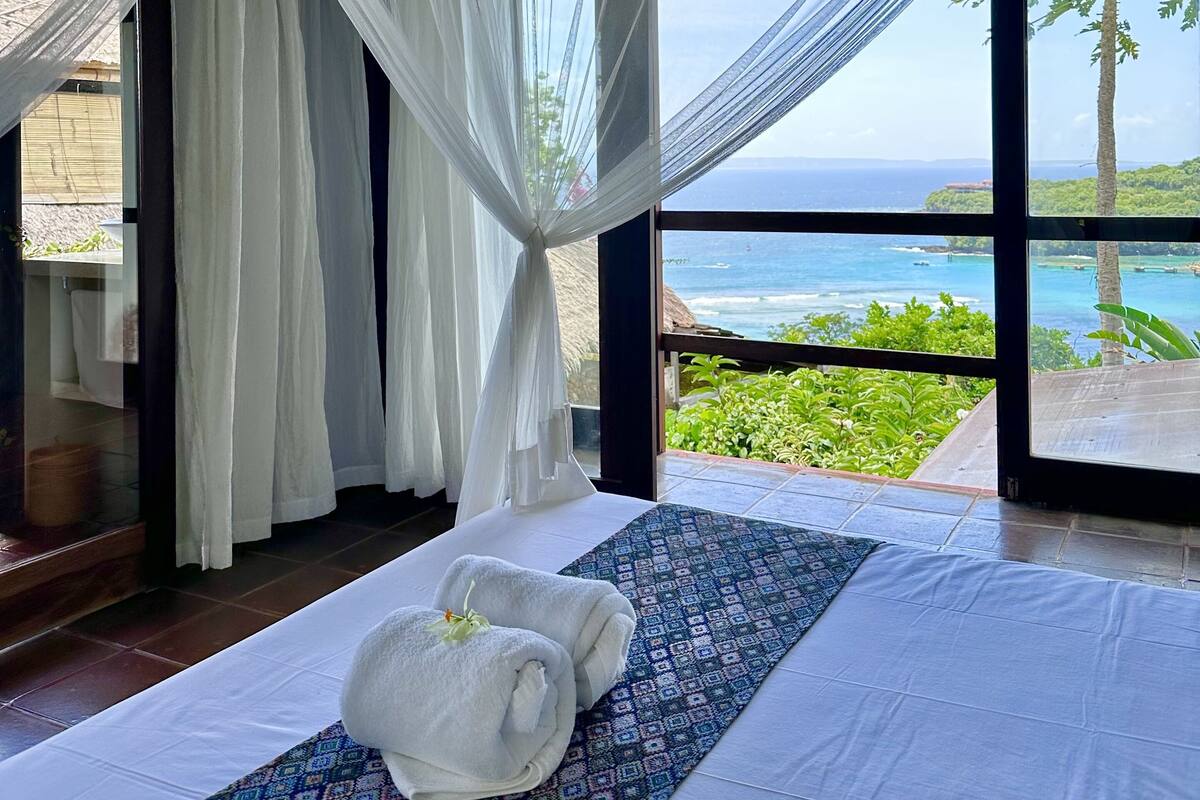 Une chambre avec un grand lit, des rideaux blancs et une vue sur l’océan.
