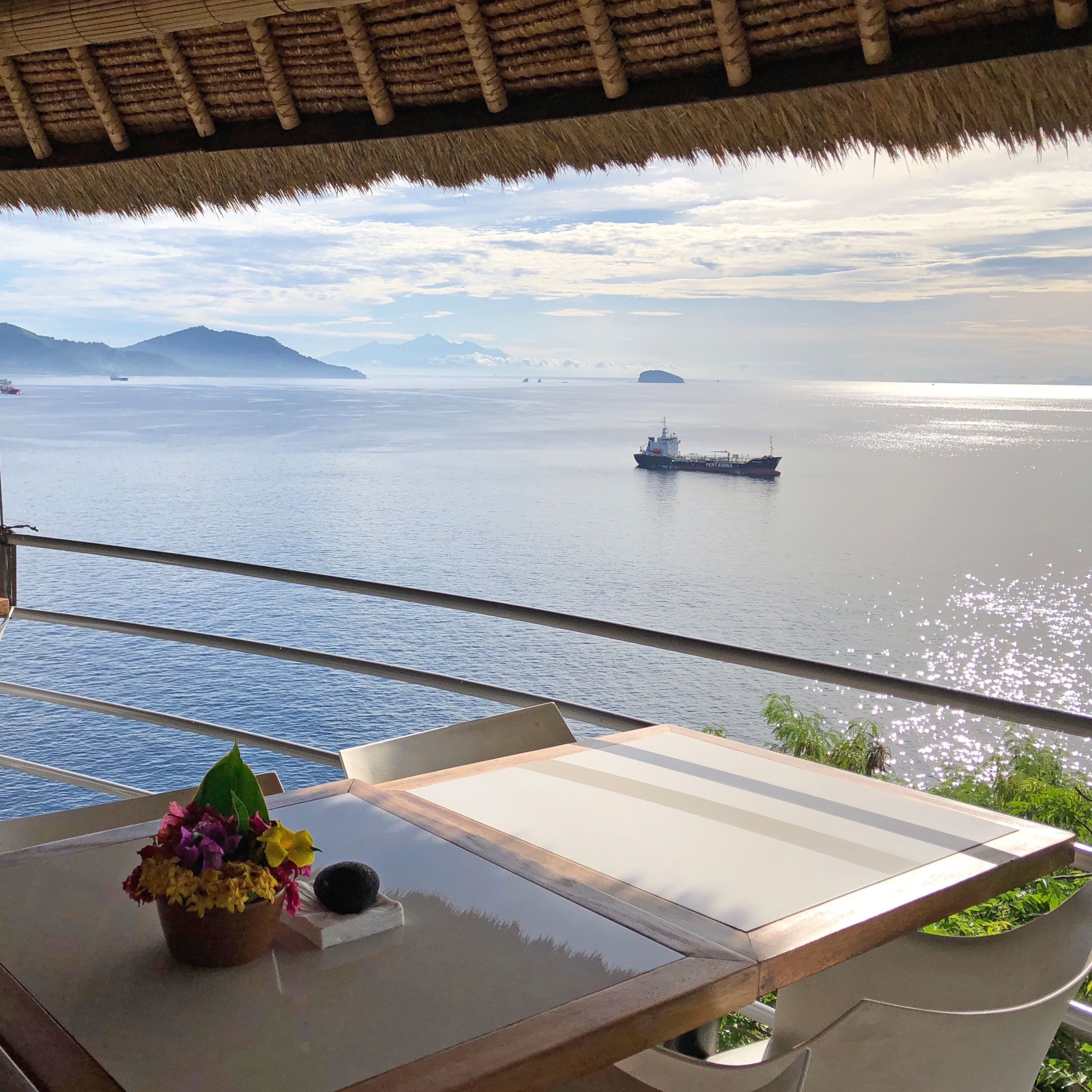 Un balcón con una mesa, una planta en maceta y vista a un mar tranquilo con un barco a lo lejos.