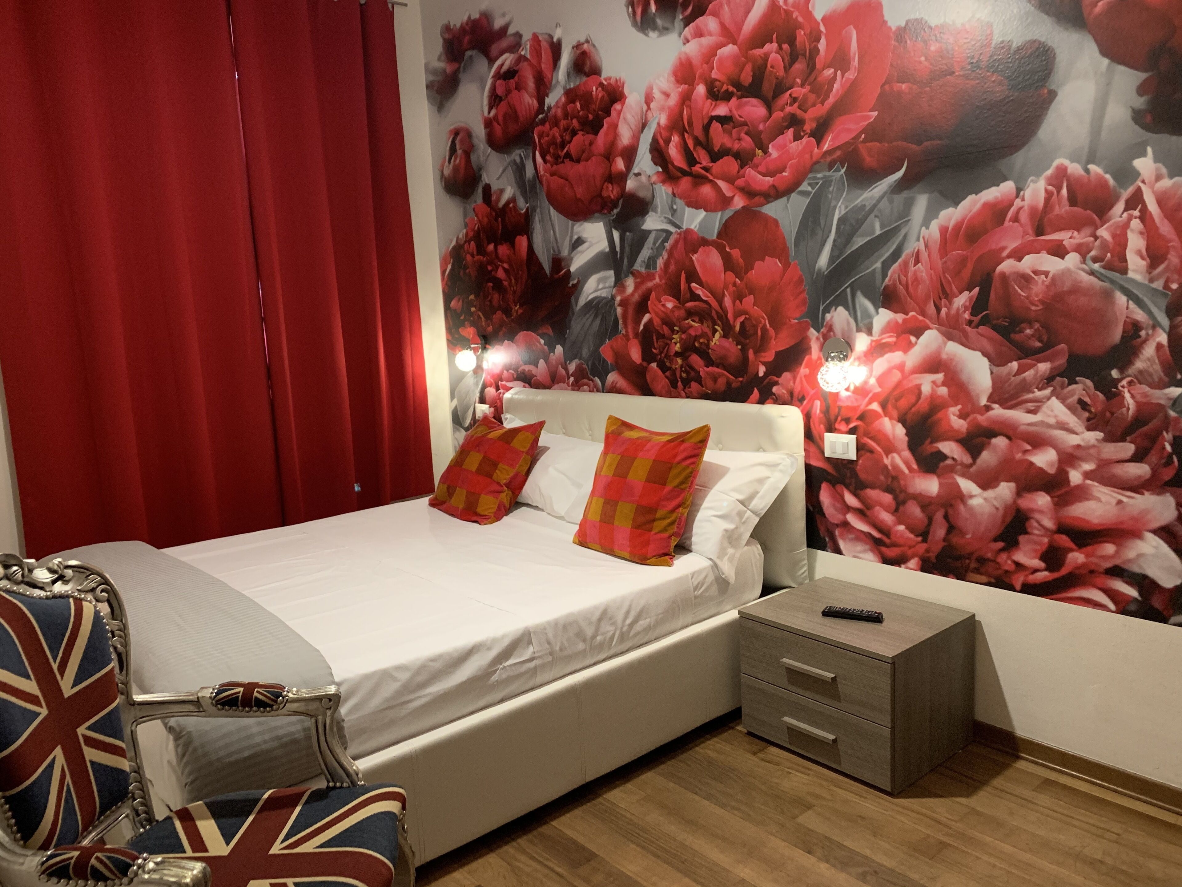 Une chambre à coucher avec un lit, des rideaux rouges, une fresque murale à motifs floraux, une table de chevet et une chaise ornée d’un motif Union Jack.