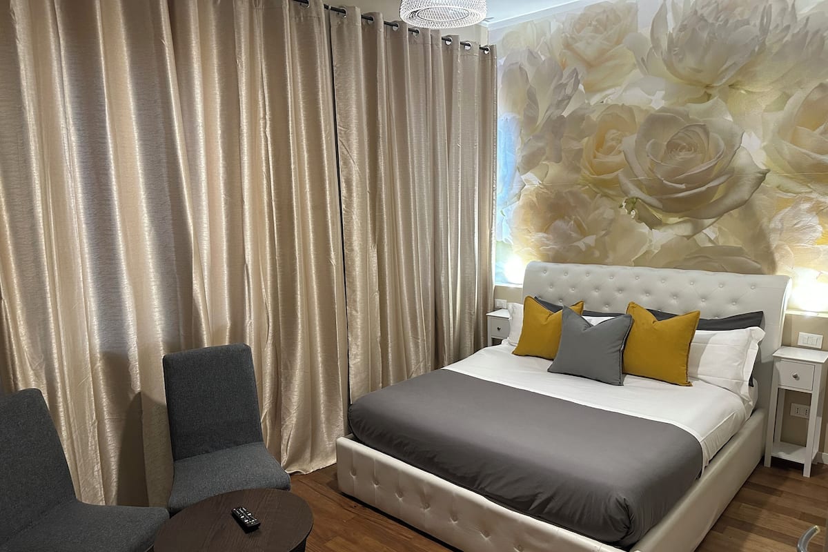 Quarto de hotel com uma cama grande, duas poltronas, uma mesinha e um mural floral na parede.