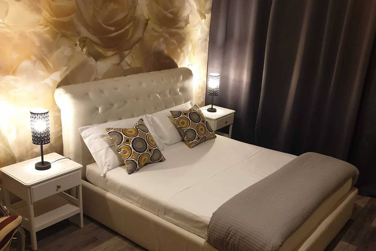 Quarto com cabeceira estofada, duas mesas de cabeceira com luminárias, cama com almofadas estampadas e papel de parede floral.