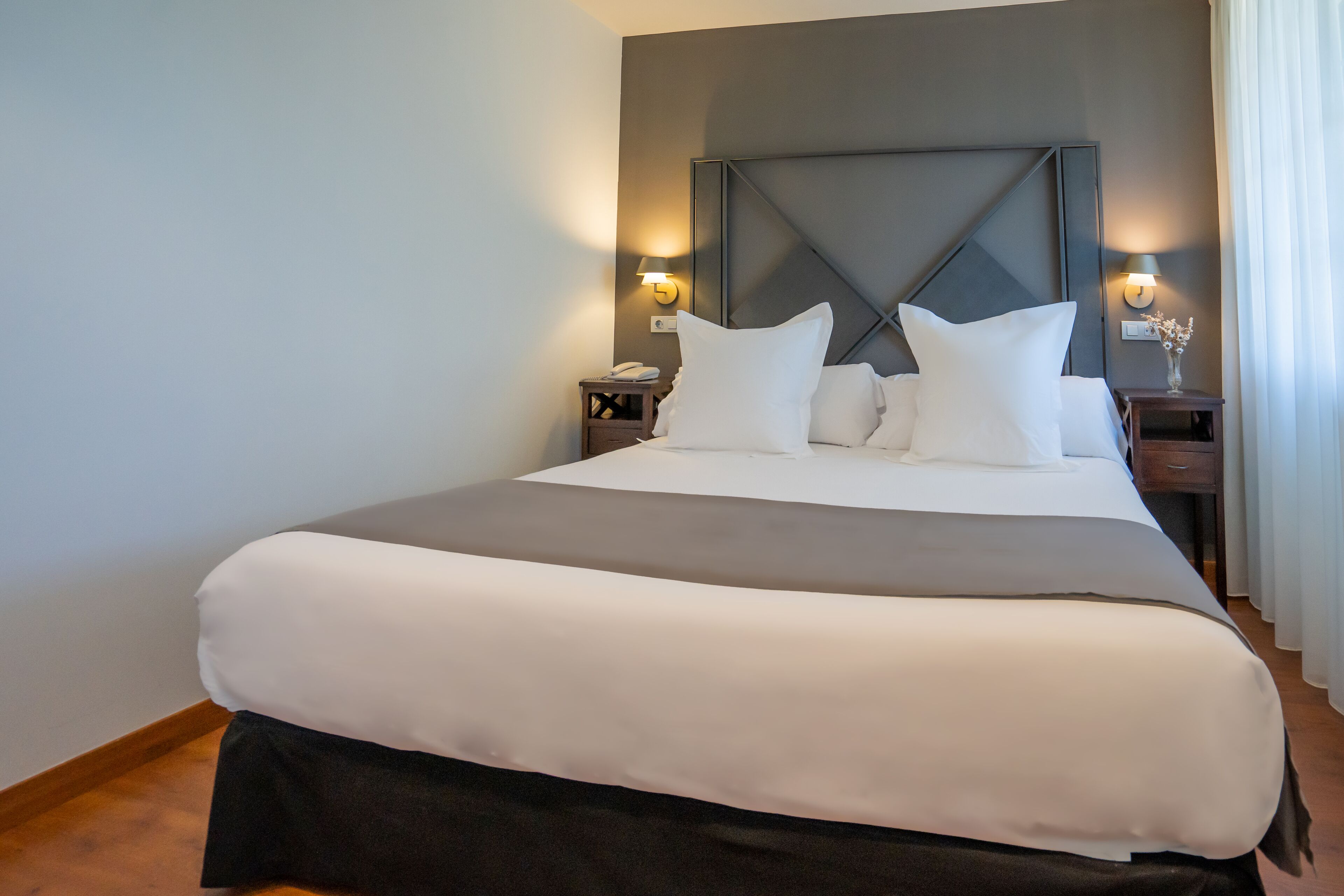 Una cama bien hecha con ropa de cama blanca y gris, un cabecero oscuro y dos mesitas de noche con lámparas.