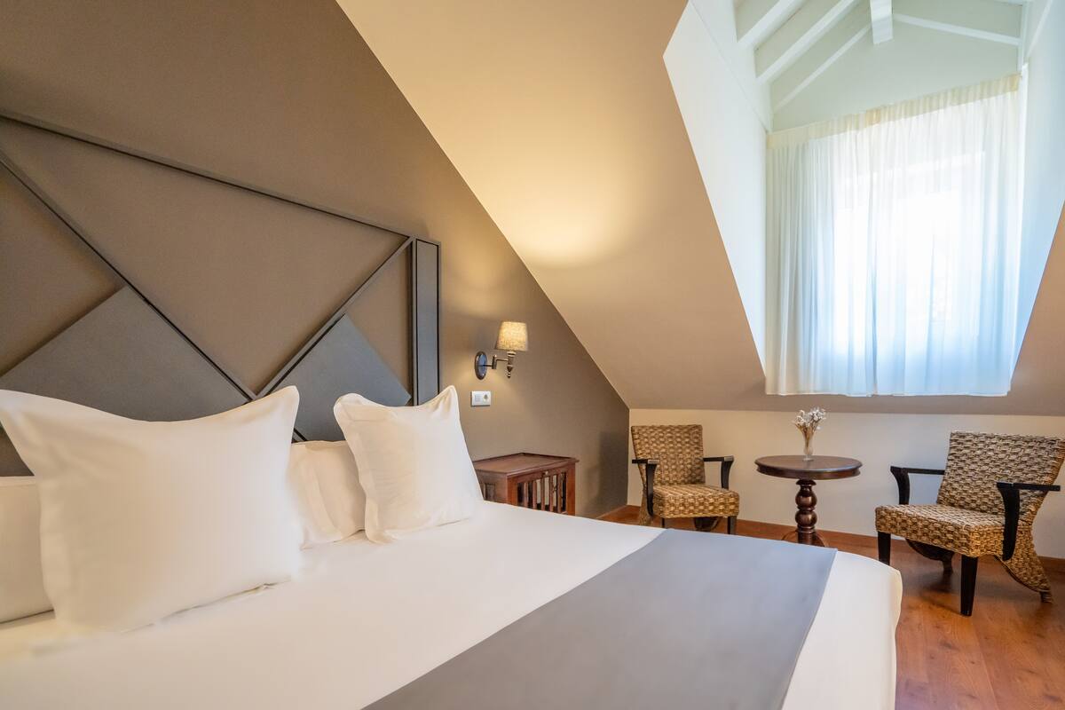 A hotel room with a large bed, two chairs, a small table, and a window with curtains.
