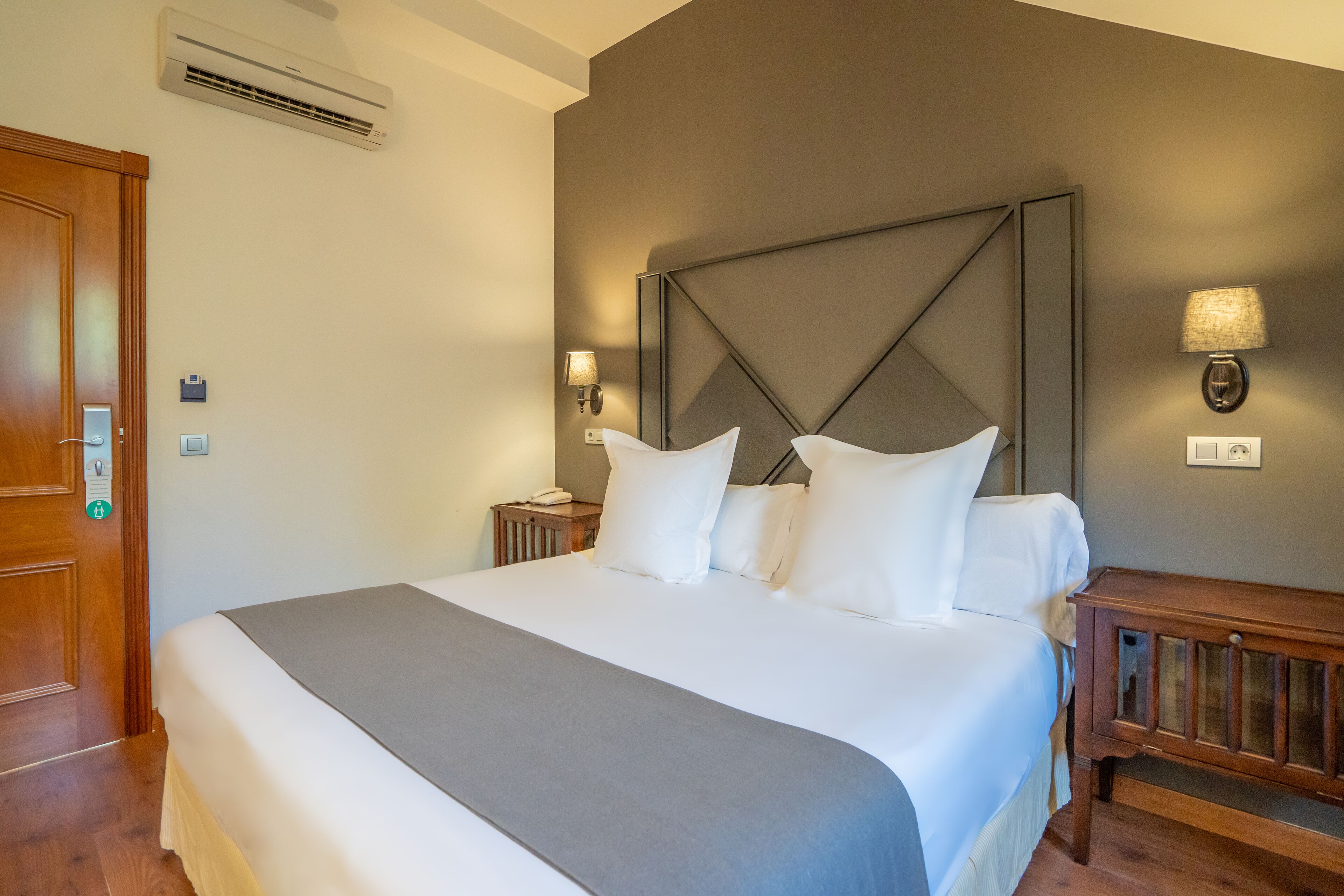 Een hotelkamer met een groot bed, een houten hoofdbord, een nachtkastje met een lamp en een airconditioning op de muur.