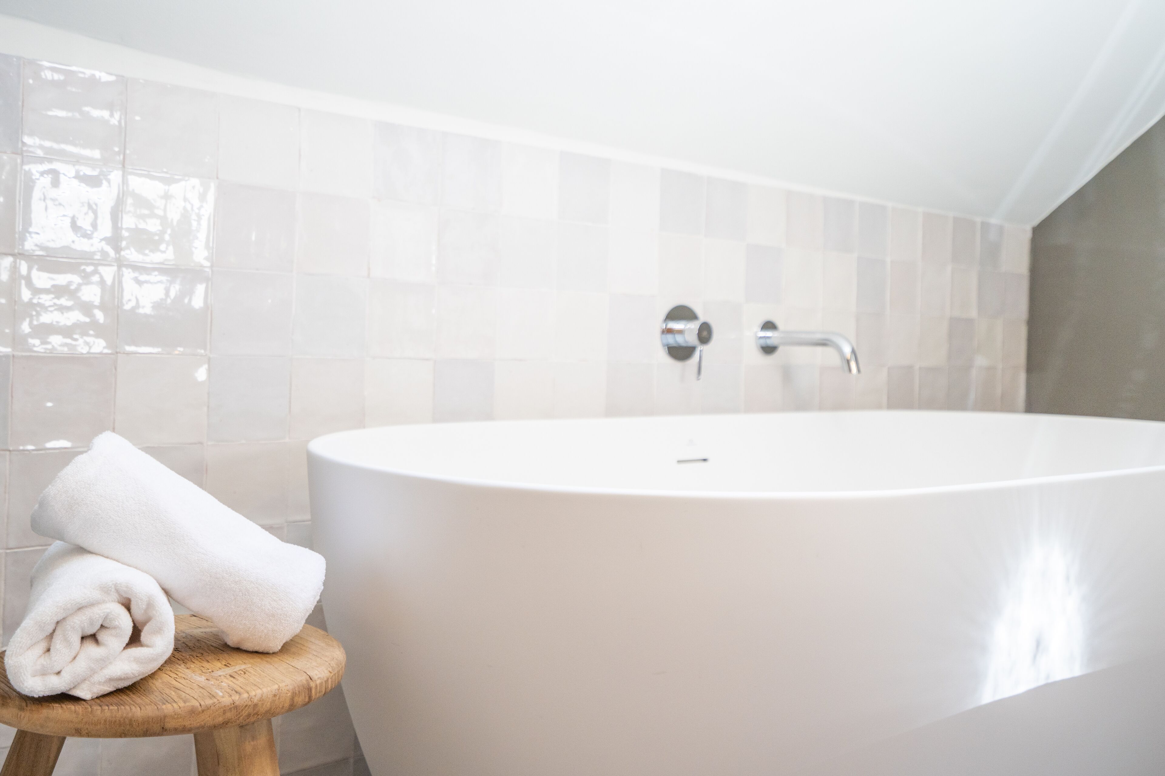 Un baño moderno con una bañera independiente, un taburete de madera y una toalla enrollada, además de paredes de azulejos.