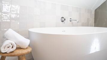 Un baño moderno con una bañera independiente, un taburete de madera y una toalla enrollada, además de paredes de azulejos.