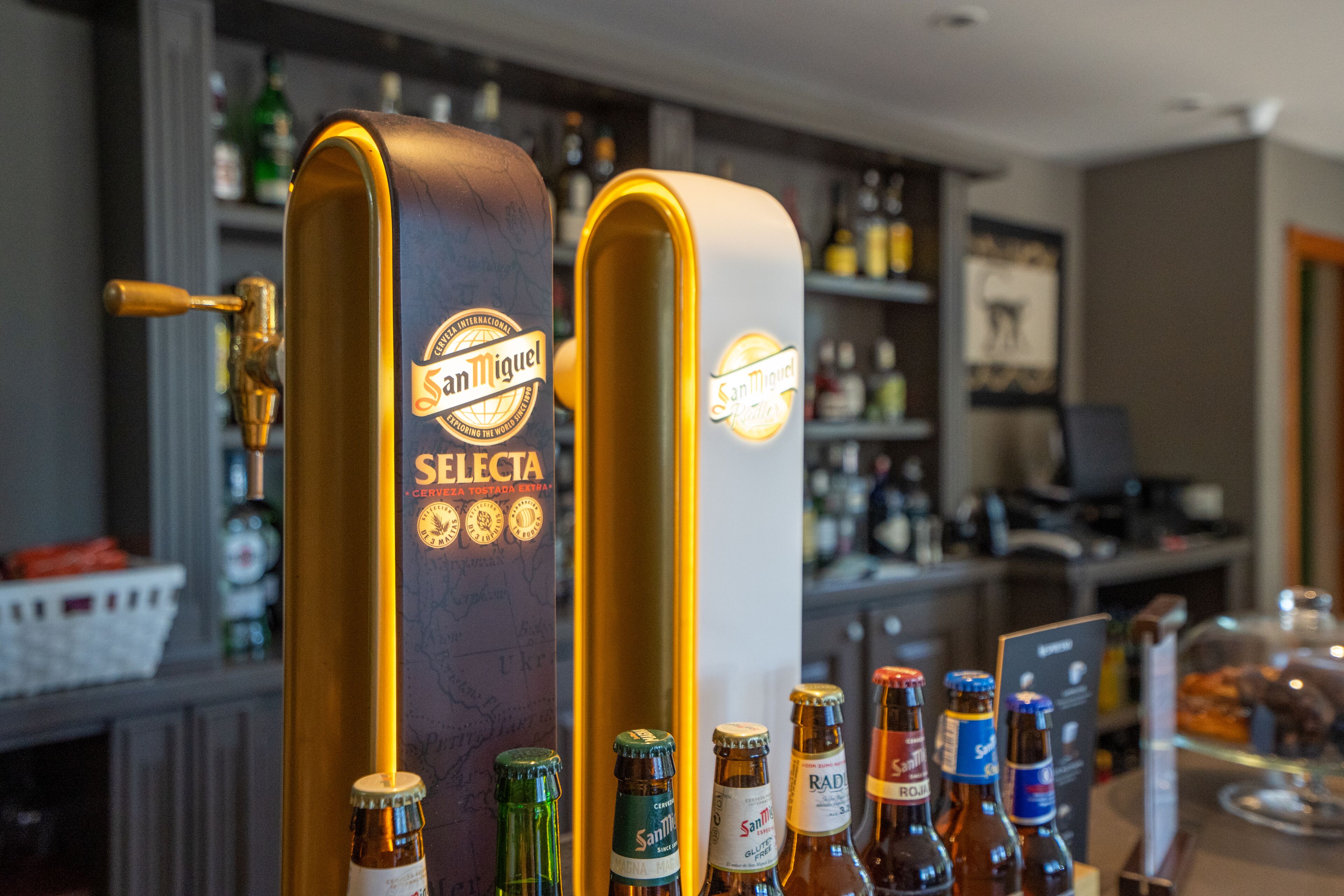 Twee biertappunten met de aanduiding 'San Miguel Selecta' in een bar, met verschillende flessen en een toonbank op de achtergrond.