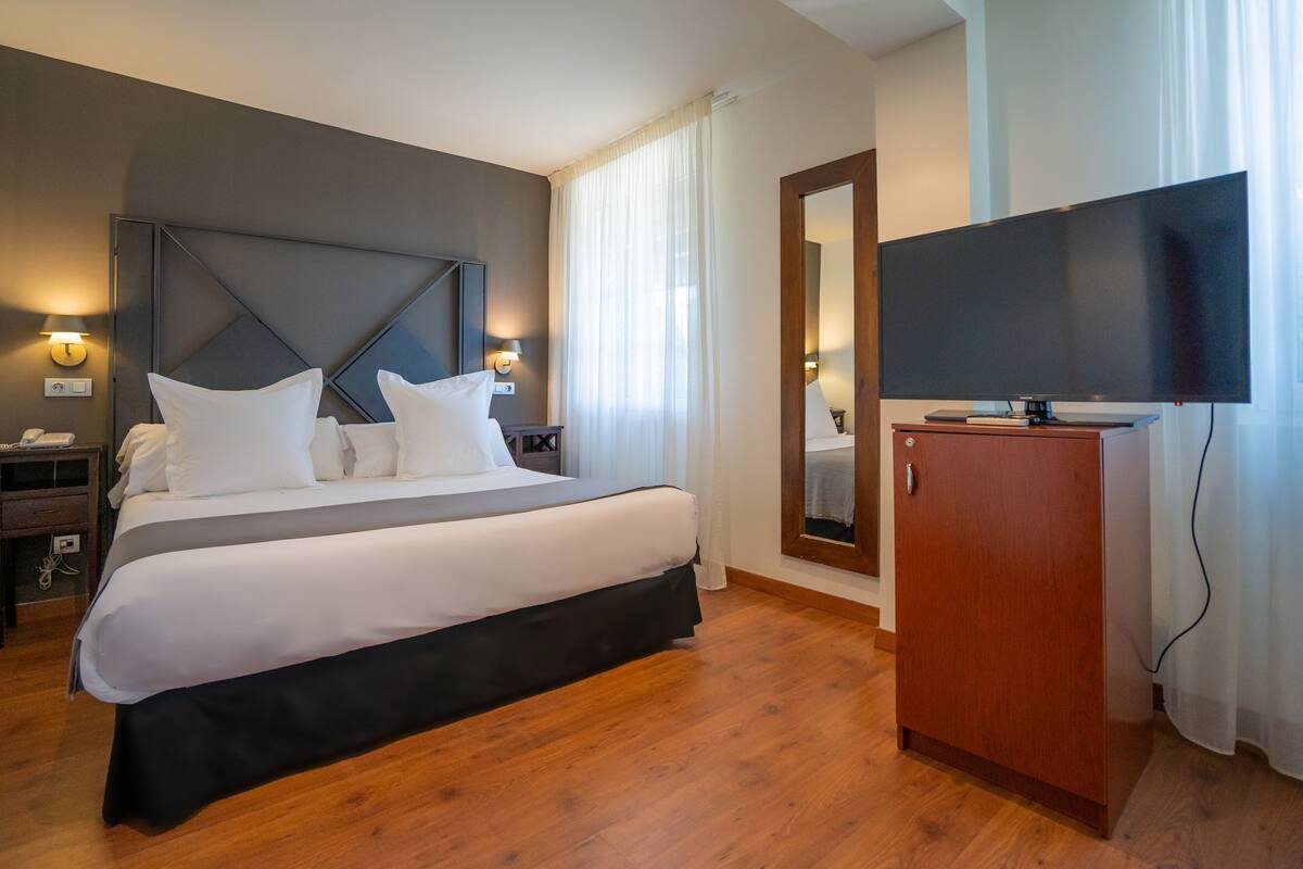 A hotel room with a large bed, a wooden wardrobe, a flat-screen TV, and a mirror.