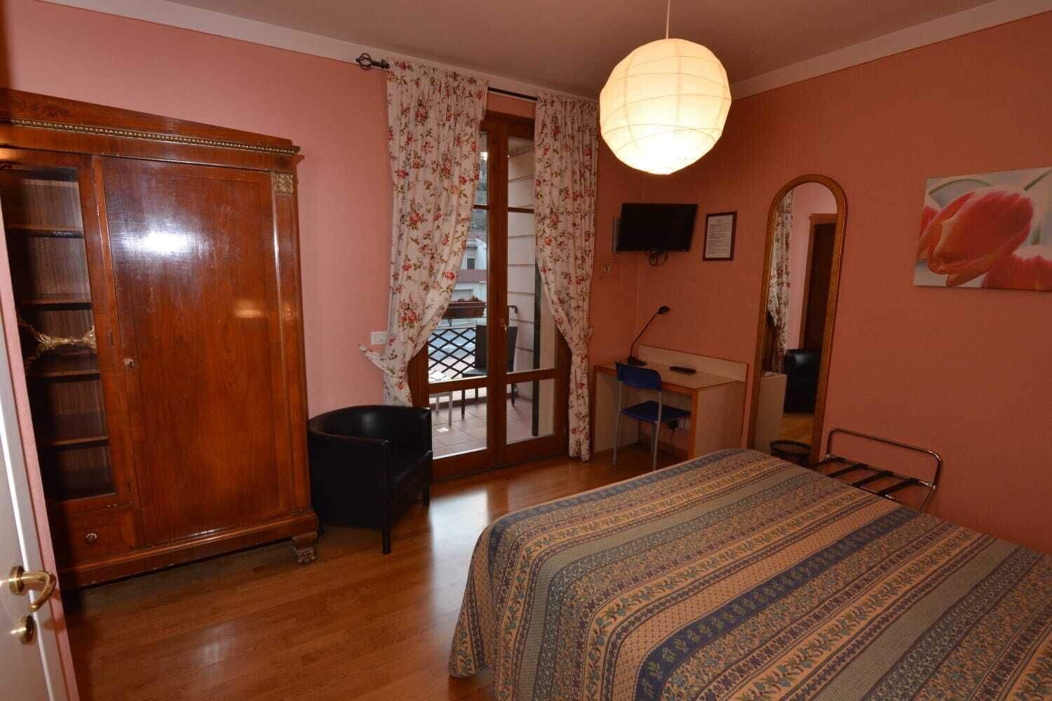 A bedroom with a wooden wardrobe, a desk, a chair, a bed with a patterned bedspread, a hanging light fixture, and a mirror on the wall.