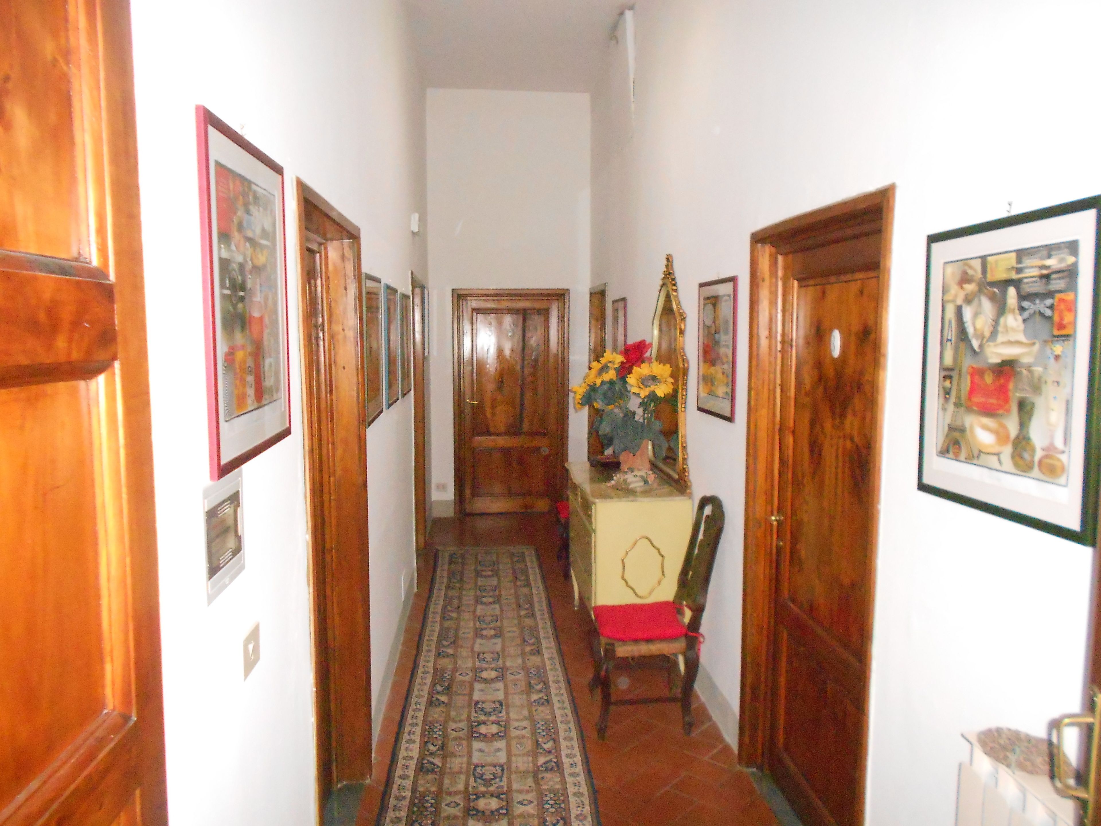 Un pasillo con puertas de madera, cuadros en las paredes, un tapete estampado y una silla con cojín rojo.