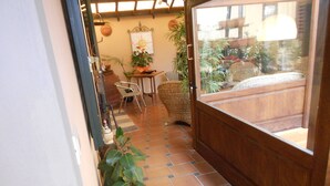 A narrow hallway with a tiled floor leading to a wooden door. There is a wicker chair and a small table with a plant on it.