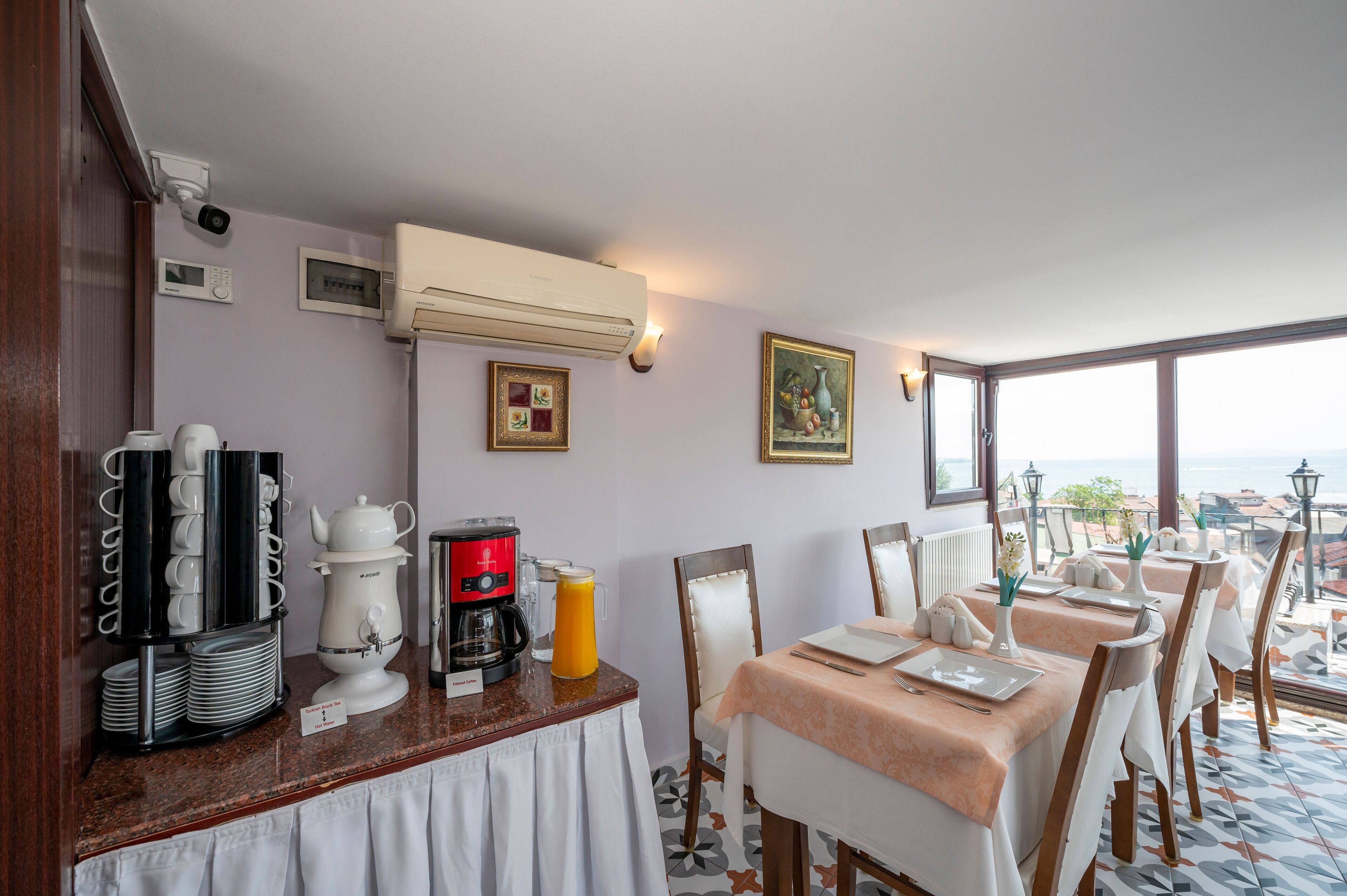 A breakfast area with a coffee machine, tea set, and fruit punch. There are tables with white tablecloths and chairs.