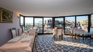 A dining area with tables and chairs, large windows with a city view, and a tiled floor.
