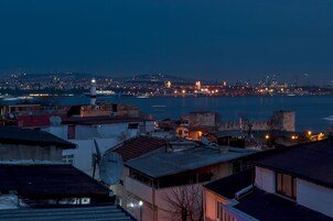 Un paisaje urbano costero al atardecer, con edificios, un faro y una estructura que parece un castillo.