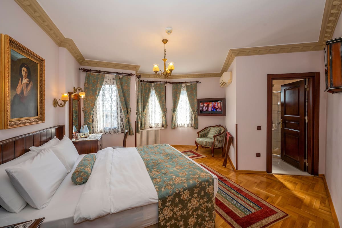 A hotel room with a large bed, a wooden headboard, a chandelier, a television, and a door leading to another room.