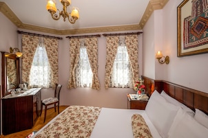 Una habitación de hotel con cama, escritorio con teléfono, silla, mesita de noche con un florero y un cuadro en la pared.