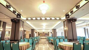 A spacious dining area with round tables and teal chairs, featuring a central chandelier and a view of the sea through large windows. - Rebecca Hotel (Zhengzhou)