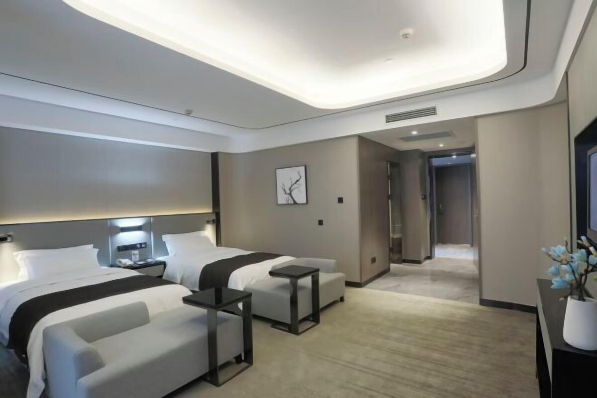 A modern hotel room with a bed, bedside tables, a sofa, a small table, and a nightstand. There is a hallway leading to another room.