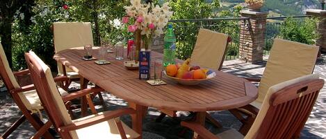 A wooden table set for six with chairs, a fruit bowl, a vase with flowers, and a bottle of Sprite.