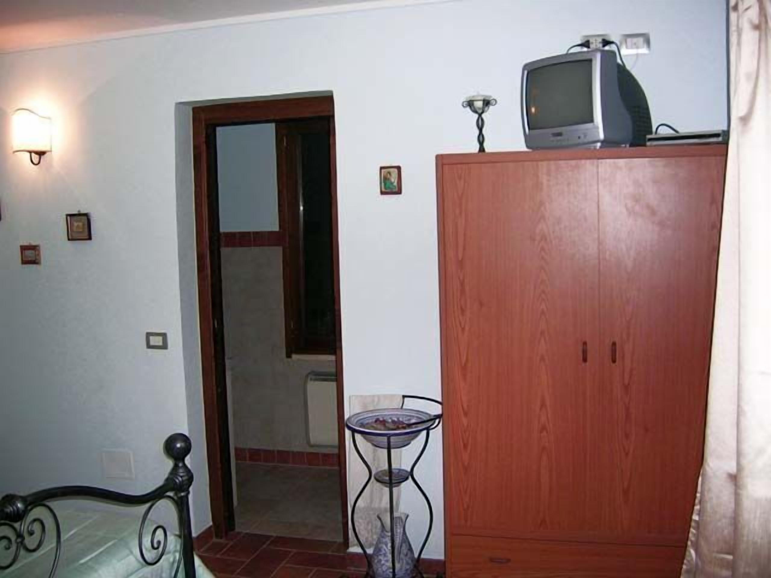 A bedroom with a wooden wardrobe, a bed with a metal frame, a small table with a vase, and a television on top of the wardrobe.
