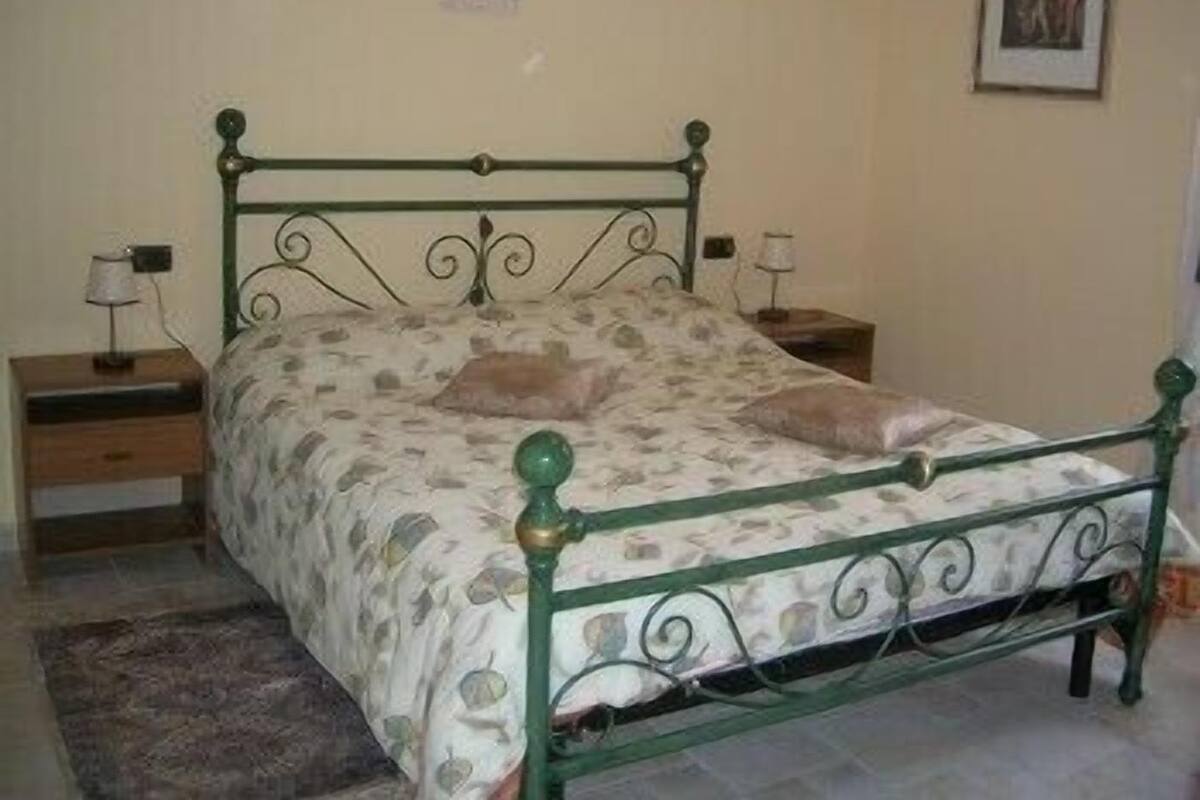 Un lit simple avec un cadre en métal vert et une couvre-lit à motifs, flanqué de deux tables de chevet sur lesquelles sont posées des lampes.