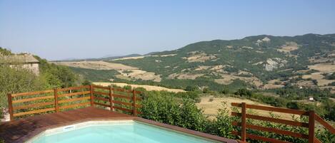 A pool with a wooden deck and metal railing, surrounded by greenery and overlooking a hilly landscape.