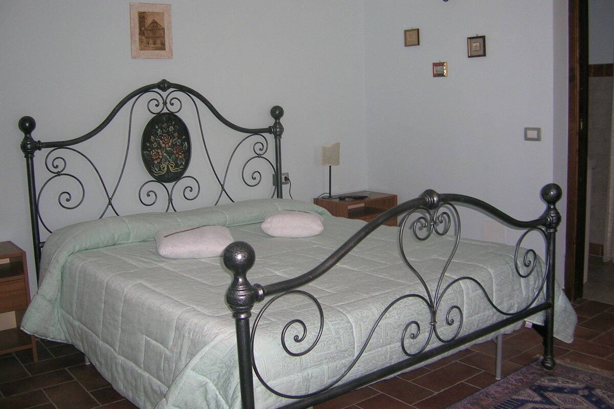 Un lit avec un cadre en métal, une couvre-lit verte et deux oreillers. Il y a des appliques murales et une petite table de chevet en bois avec une lampe.