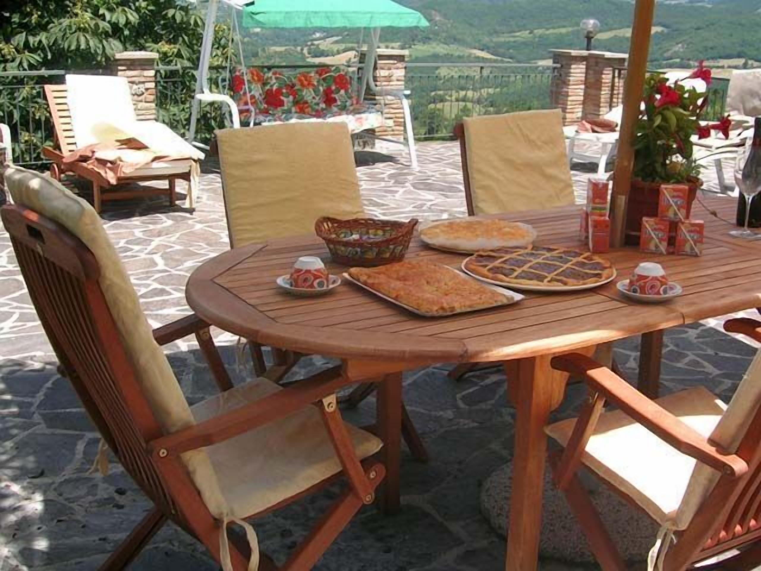 An outdoor dining area with a round wooden table, cushioned chairs, and a view of the countryside.