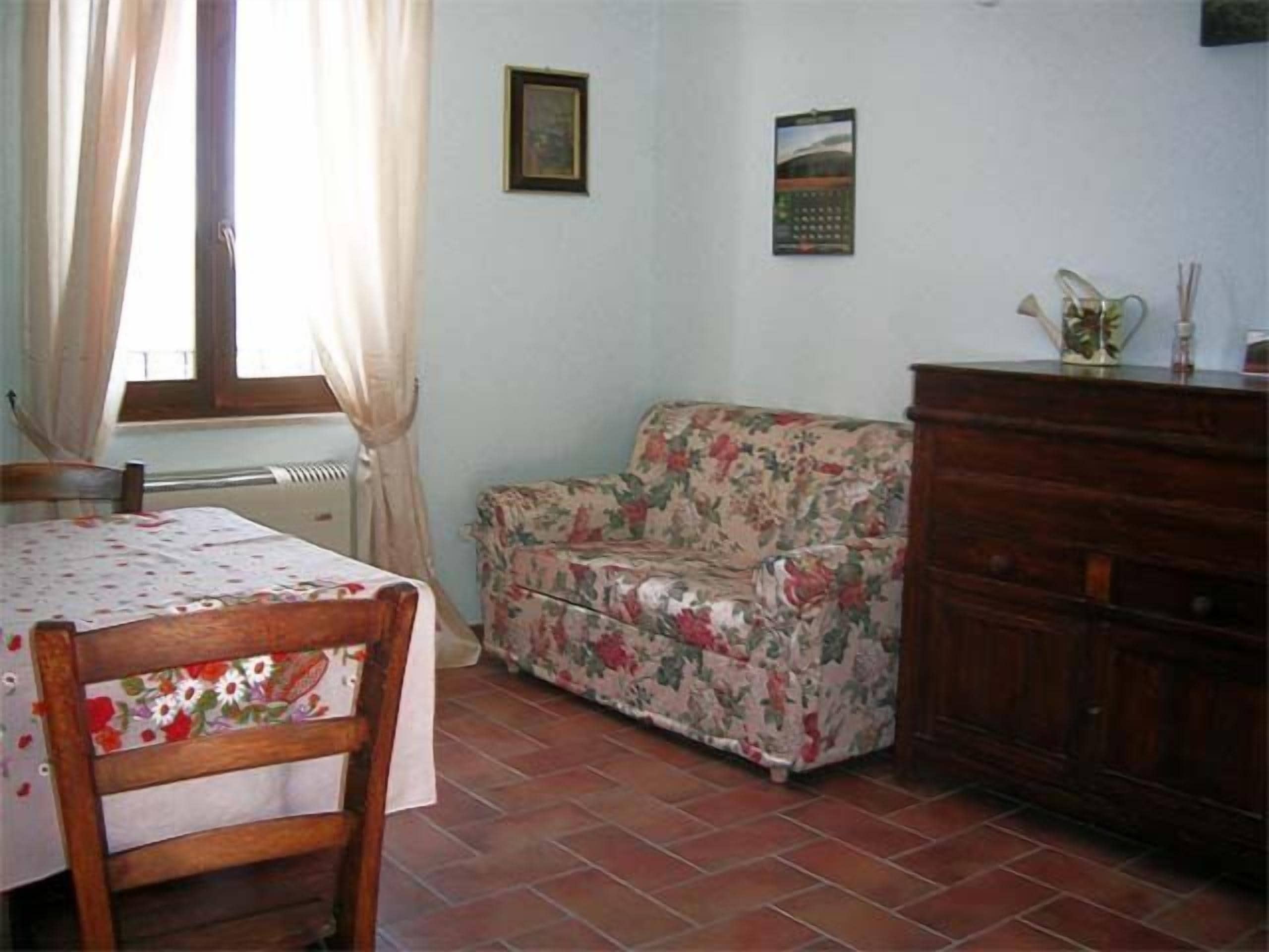 Une pièce comprenant une chaise en bois, un canapé à motifs floraux, une commode en bois et une fenêtre avec des rideaux.