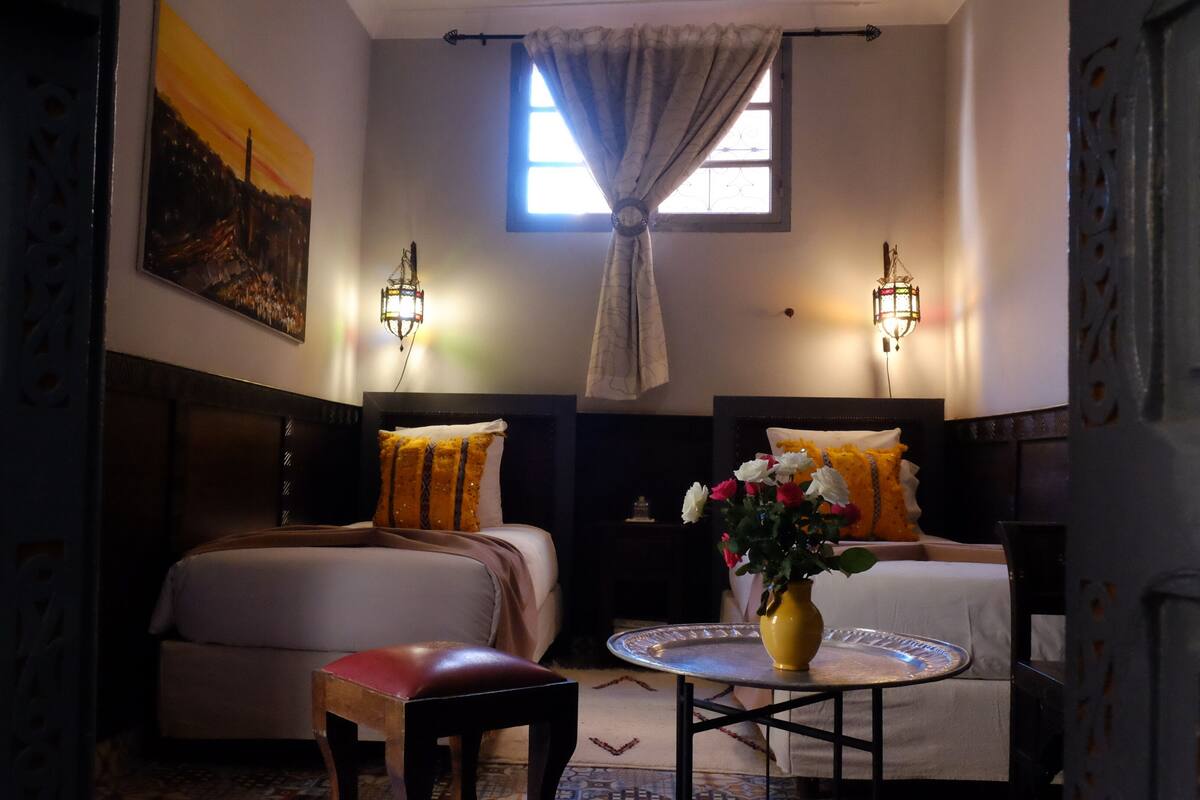 Ein Hotelzimmer mit zwei Betten, einem kleinen runden Tisch mit Blumenvase, einem Sessel und einem Bild an der Wand.