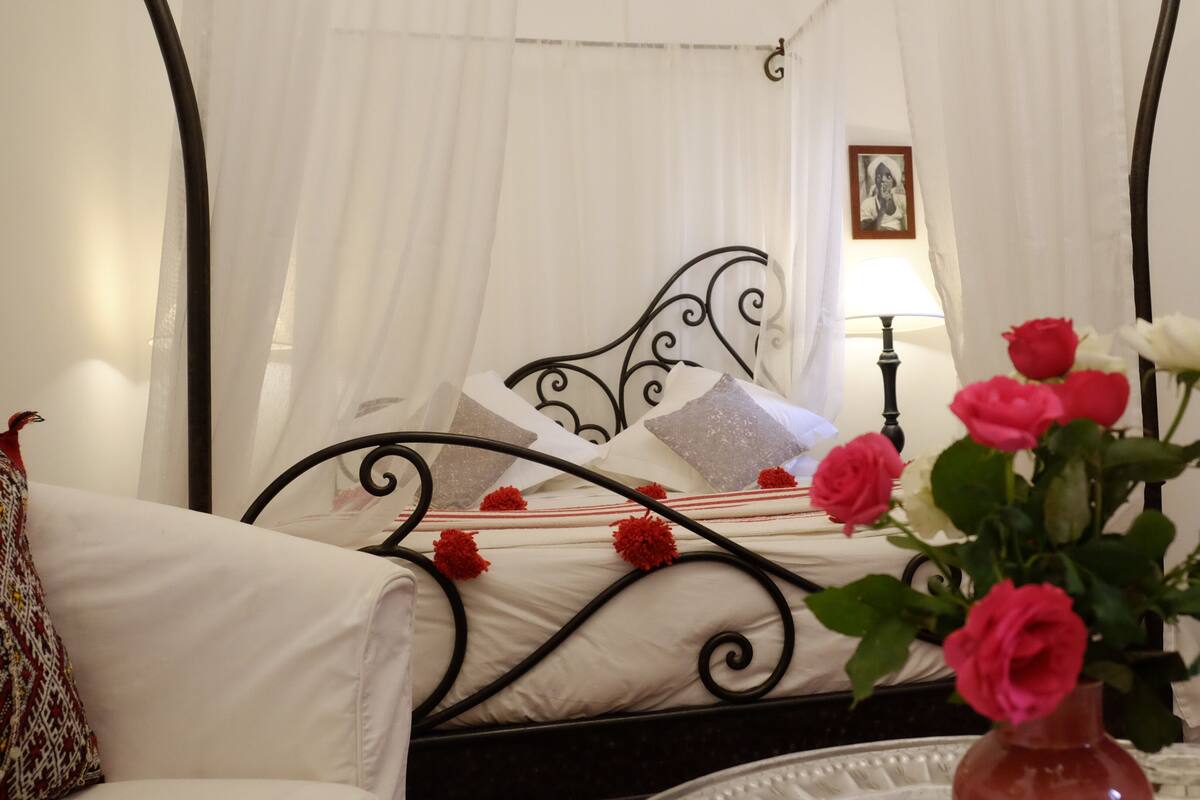 Ein Schlafzimmer mit einem Bett, weißen Vorhängen, einem Nachttisch mit Blumen und einer Lampe.