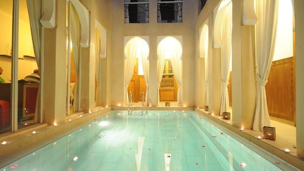 Una piscina interior con superficie de agua cristalina, rodeada de columnas y arcos, e iluminada con cálida luz ambiental.