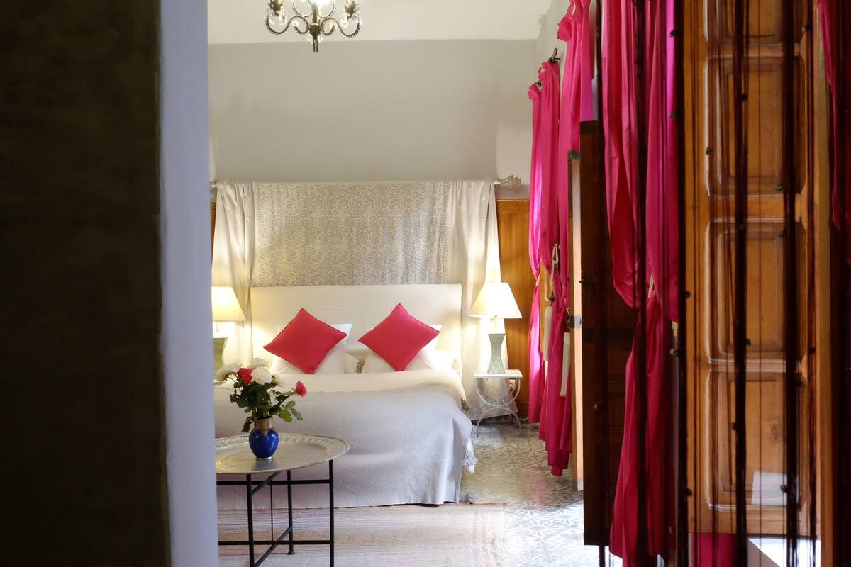 Ein Hotelzimmer mit einem Bett, zwei roten Kissen, einem Nachttisch mit Lampe und einer Vase mit Blumen.