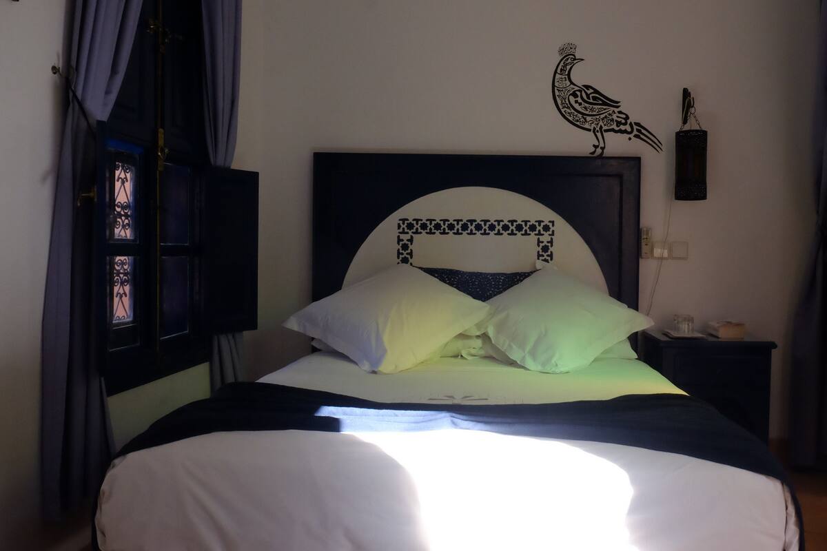 Ein Bett mit weißer Bettwäsche, ein Vorhang mit dekorativem Muster, ein Wandbild eines Vogels und ein Nachttisch mit Lampe.