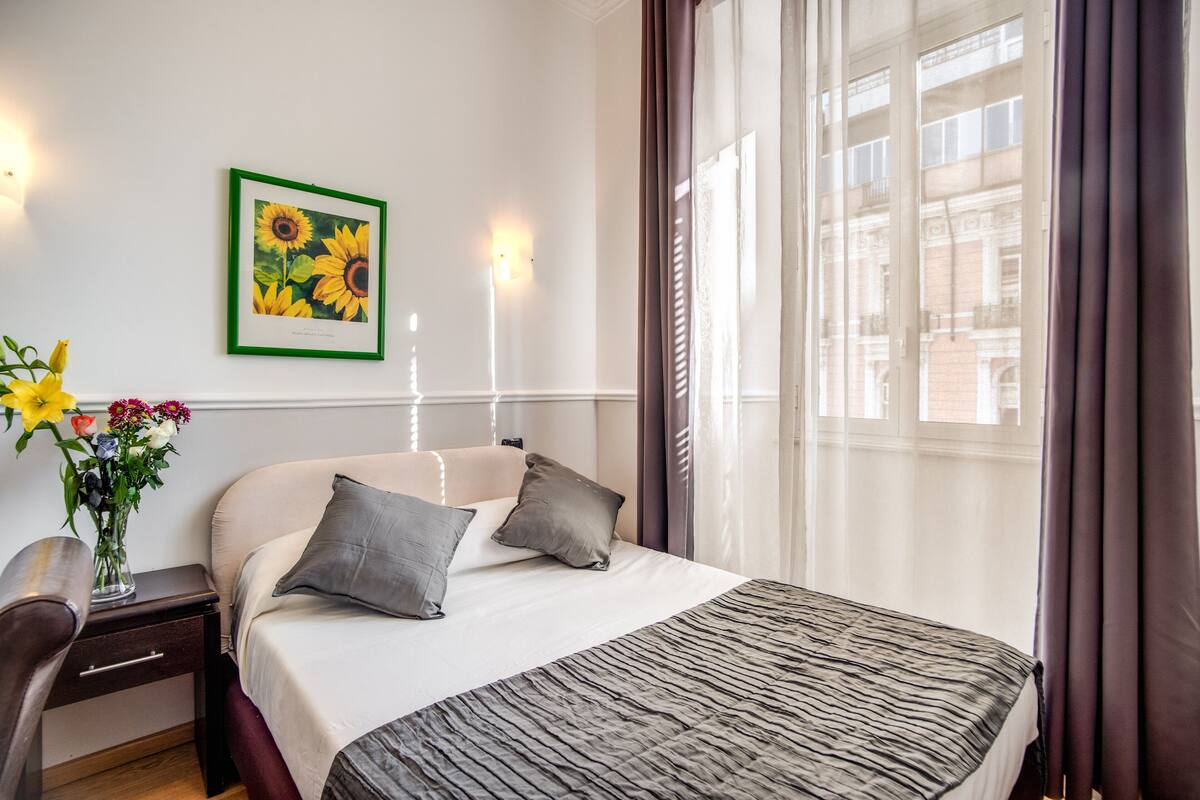A hotel room with a bed, a nightstand, a desk, a chair, a vase with flowers, and a framed picture on the wall.