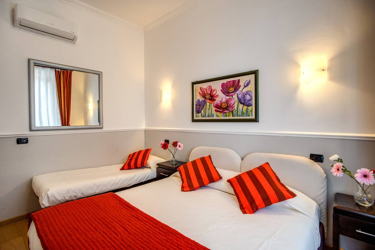 A hotel room with two beds, each with white bedding and red and black striped pillows. There is a painting of flowers on the wall, a vase with flowers on the nightstand, and an air conditioning unit above the beds.