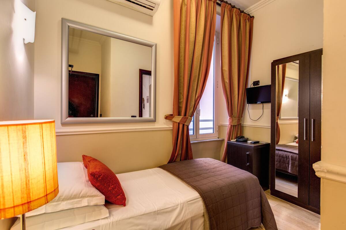 A hotel room with a bed, a bedside table with a lamp, a dresser, a mirror, and a TV on the wall.