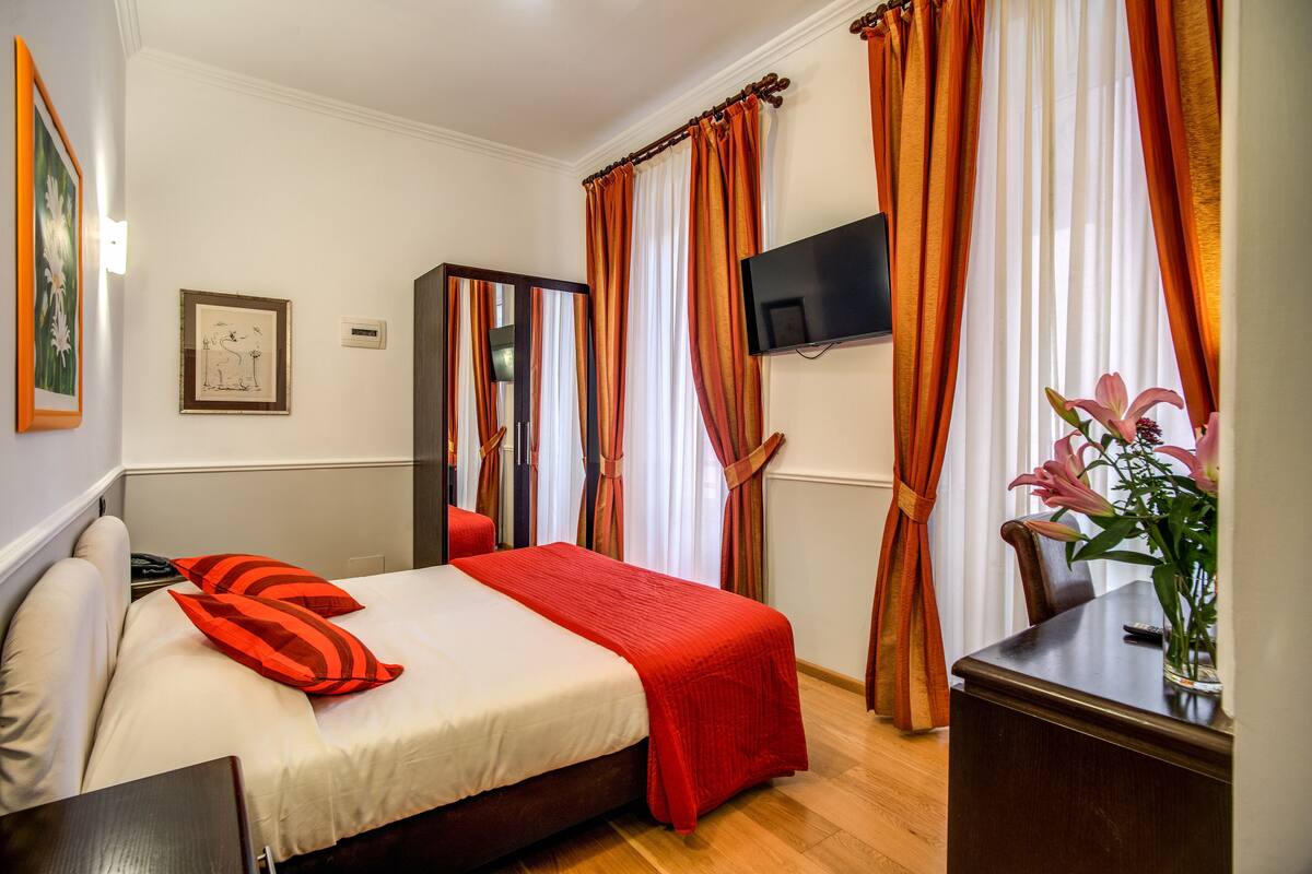 A hotel room with a bed, a TV mounted on the wall, a desk with a vase of flowers, and a window with curtains.