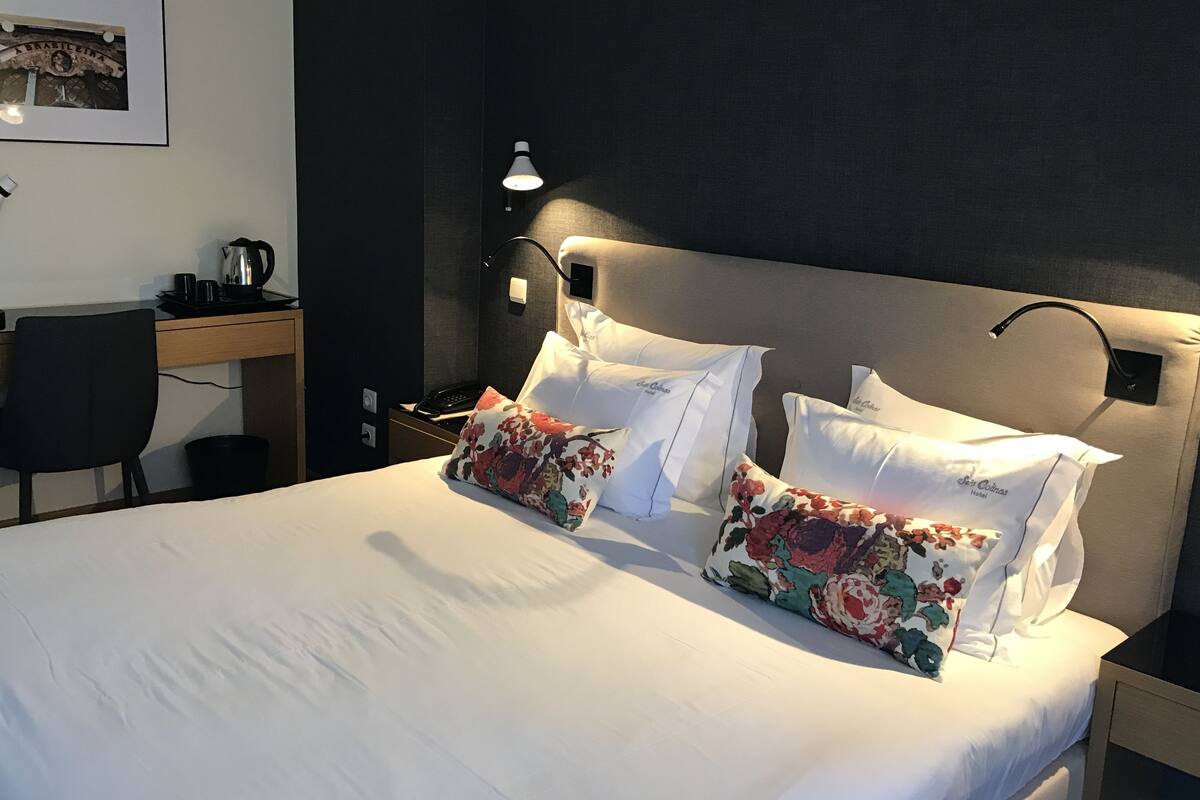 A neatly made bed with floral pillows, a bedside lamp, and a desk with a telephone and a coffee maker.