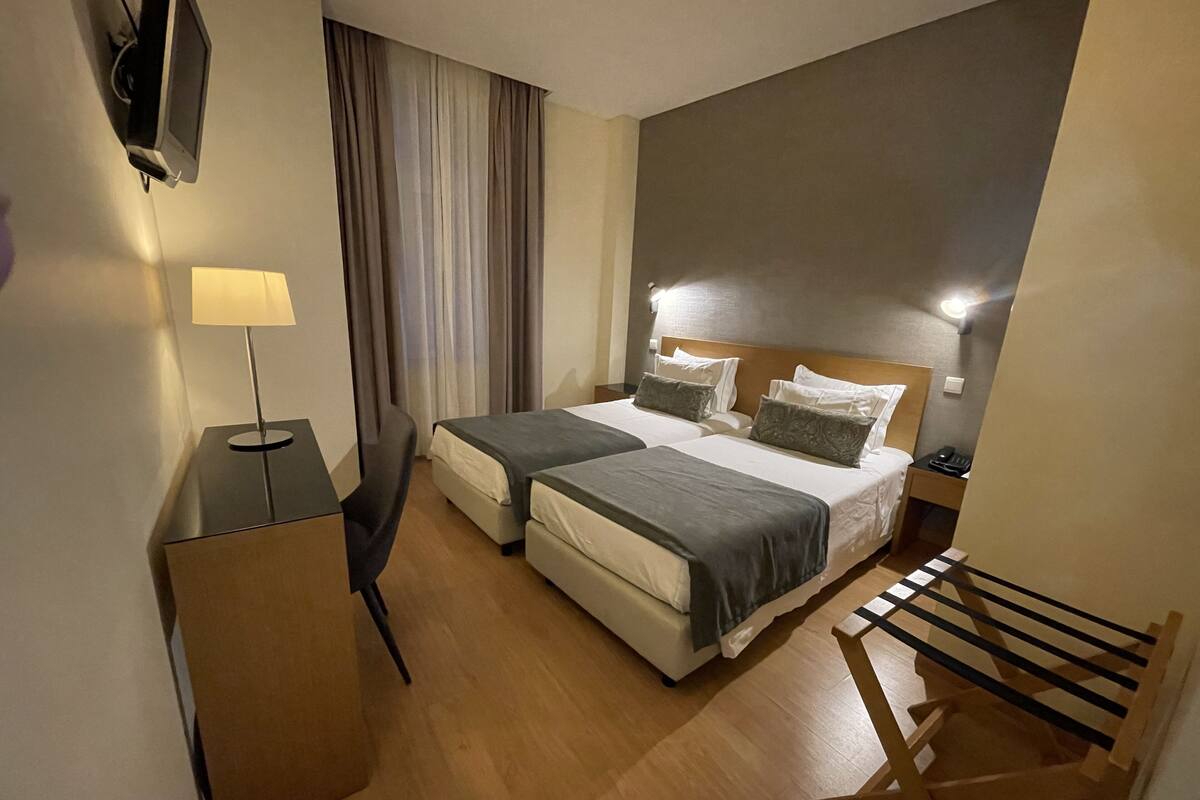 A hotel room with two beds, a desk, a chair, a lamp, and a TV.